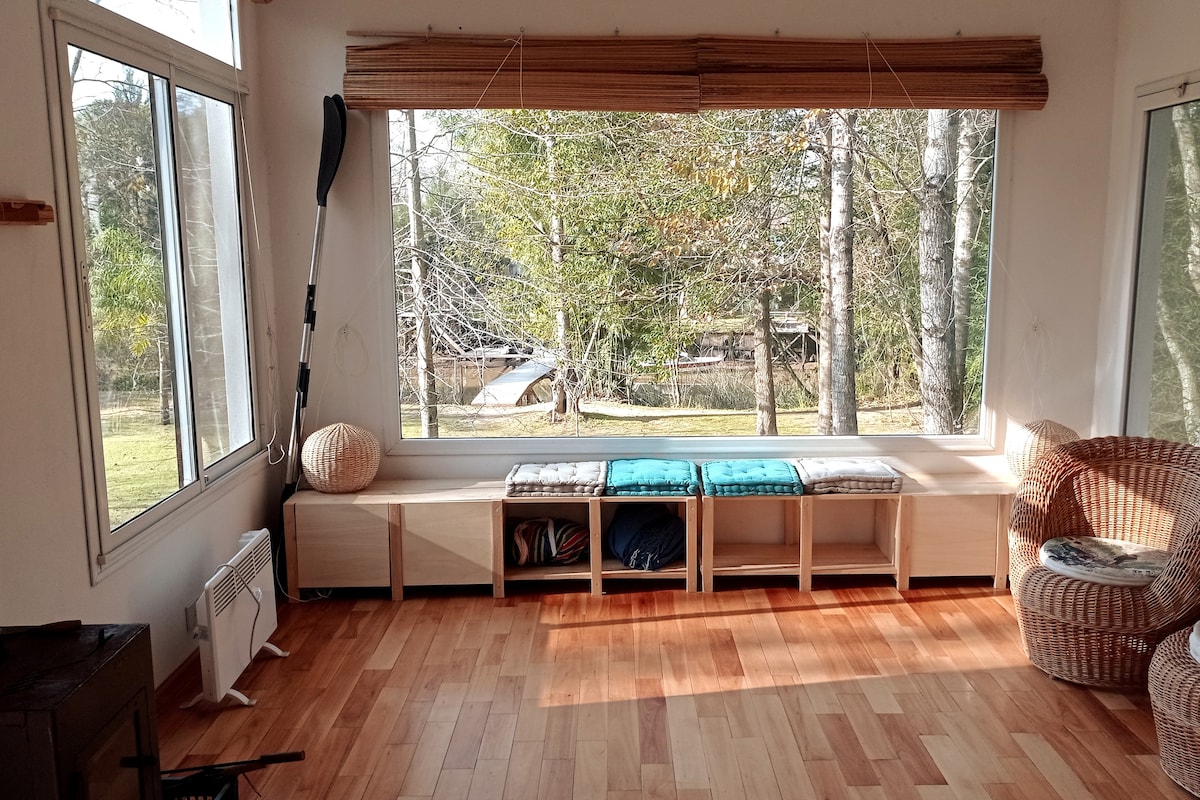 A bright living space is enhanced by large windows showcasing views of trees and the outdoors. A wooden bench is lined with cushions, and storage is provided below. Natural light illuminates the wooden flooring, creating a serene environment.