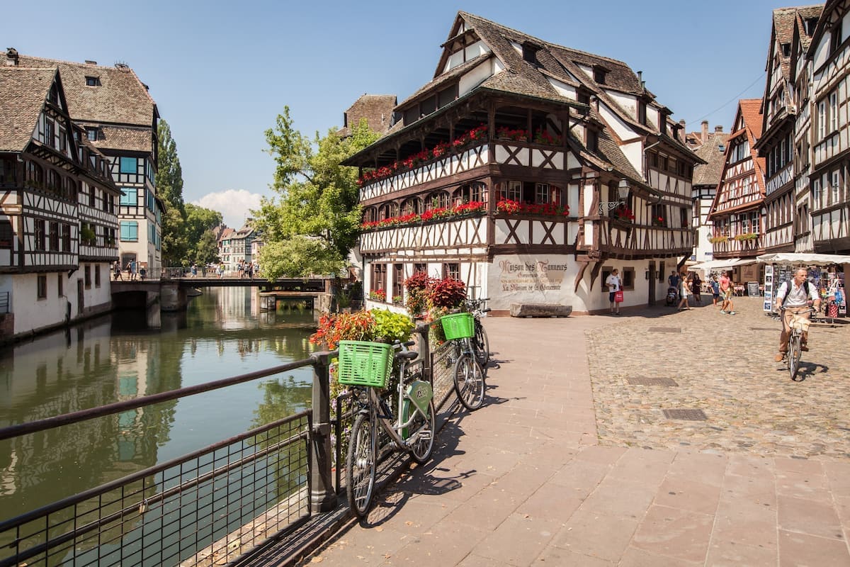 A historic area showcases traditional half-timbered houses alongside a peaceful waterway. Green bicycles are parked near a cobblestone square adorned with potted plants, inviting exploration. The scene is enhanced by lush greenery and reflections of the architecture on the calm surface of the water.