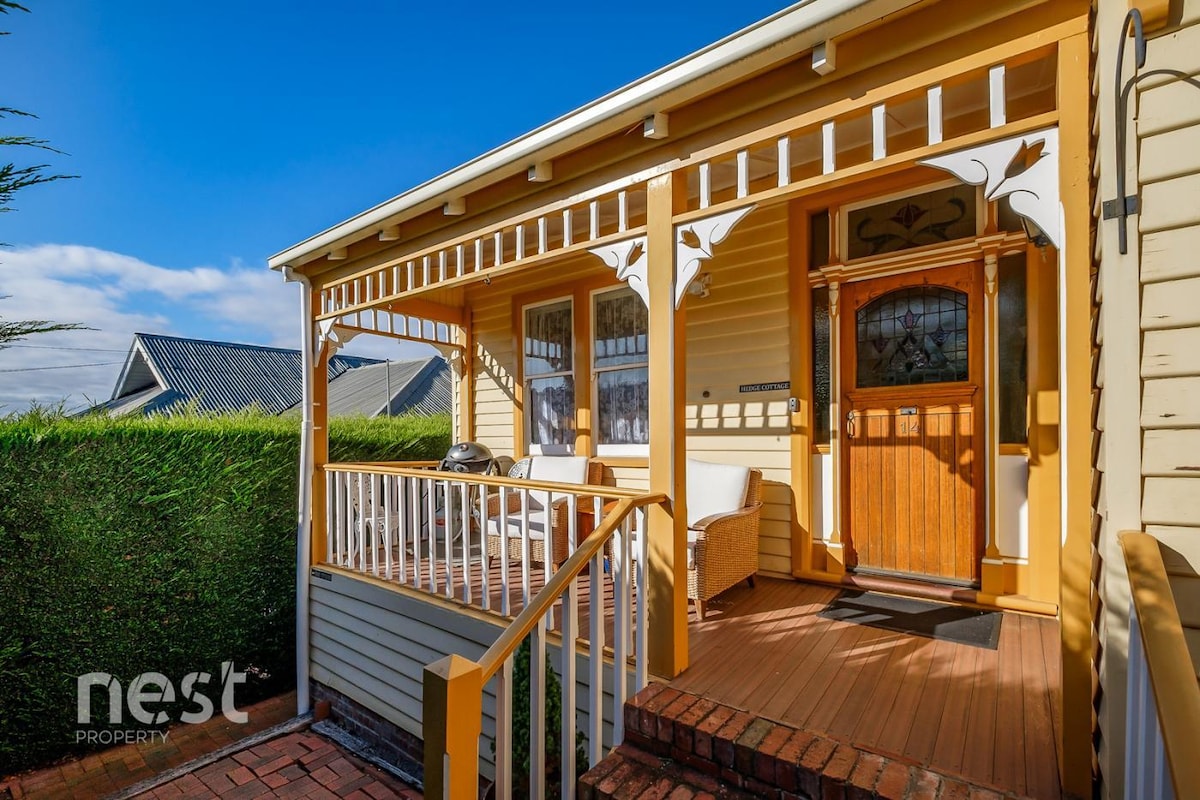 A charming entryway is framed by decorative white railings and vibrant yellow walls. Two comfortable chairs are positioned on the porch, inviting relaxation. A wooden front door features intricate glass detailing, enhancing the welcoming appeal. Lush greenery surrounds the entrance, offering a sense of privacy.