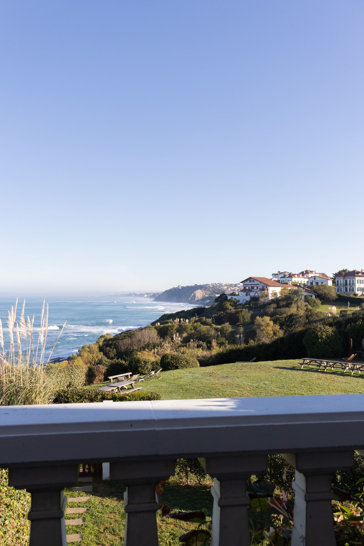 A coastal landscape is visible, featuring rolling hills and a sandy beach with gentle waves. The scene includes a grassy area with scenic seating and overlooks charming houses along the coastline under a clear blue sky.
