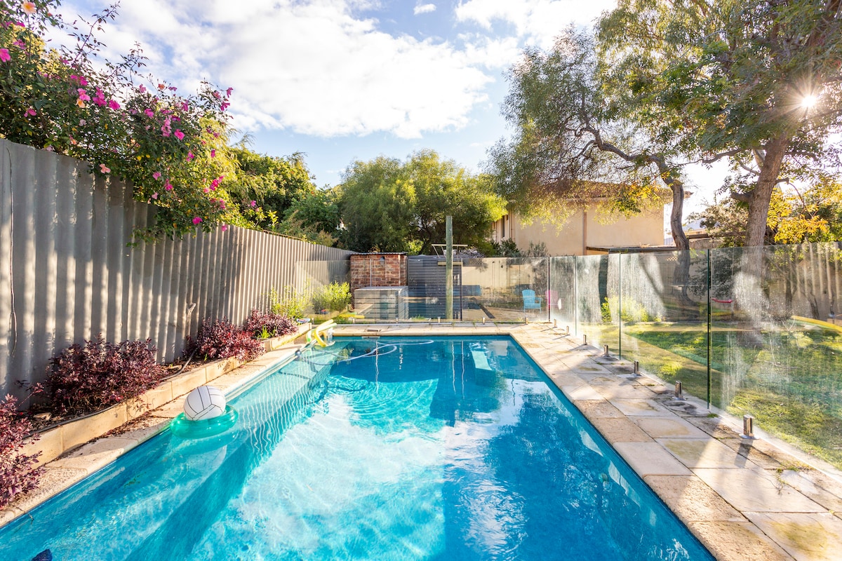 A newly designed swimming pool is framed by a landscaped garden featuring lush greenery and flowering plants. The pool area is enclosed by transparent glass fencing, enhancing safety and visibility. Sunlight reflects off the clear water, creating a refreshing ambiance.