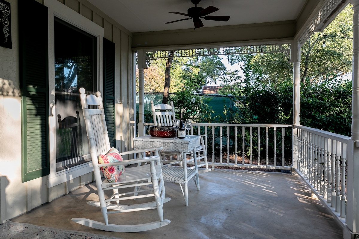 The front porch features two rocking chairs and a white wicker table, creating a casual seating area. The space is framed by greenery, providing a sense of privacy and tranquility. A ceiling fan is visible above, along with natural light filtering through the surrounding trees.