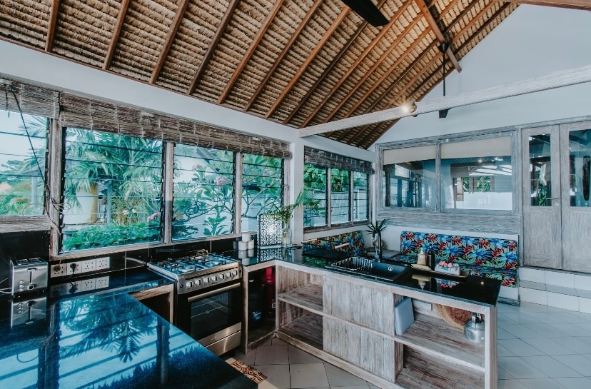The kitchen area features a modern gas stove and a full refrigerator, surrounded by large windows that provide ample natural light. A vibrant, cushioned seating area with floral patterns complements the spacious layout, creating a welcoming space for cooking and dining.
