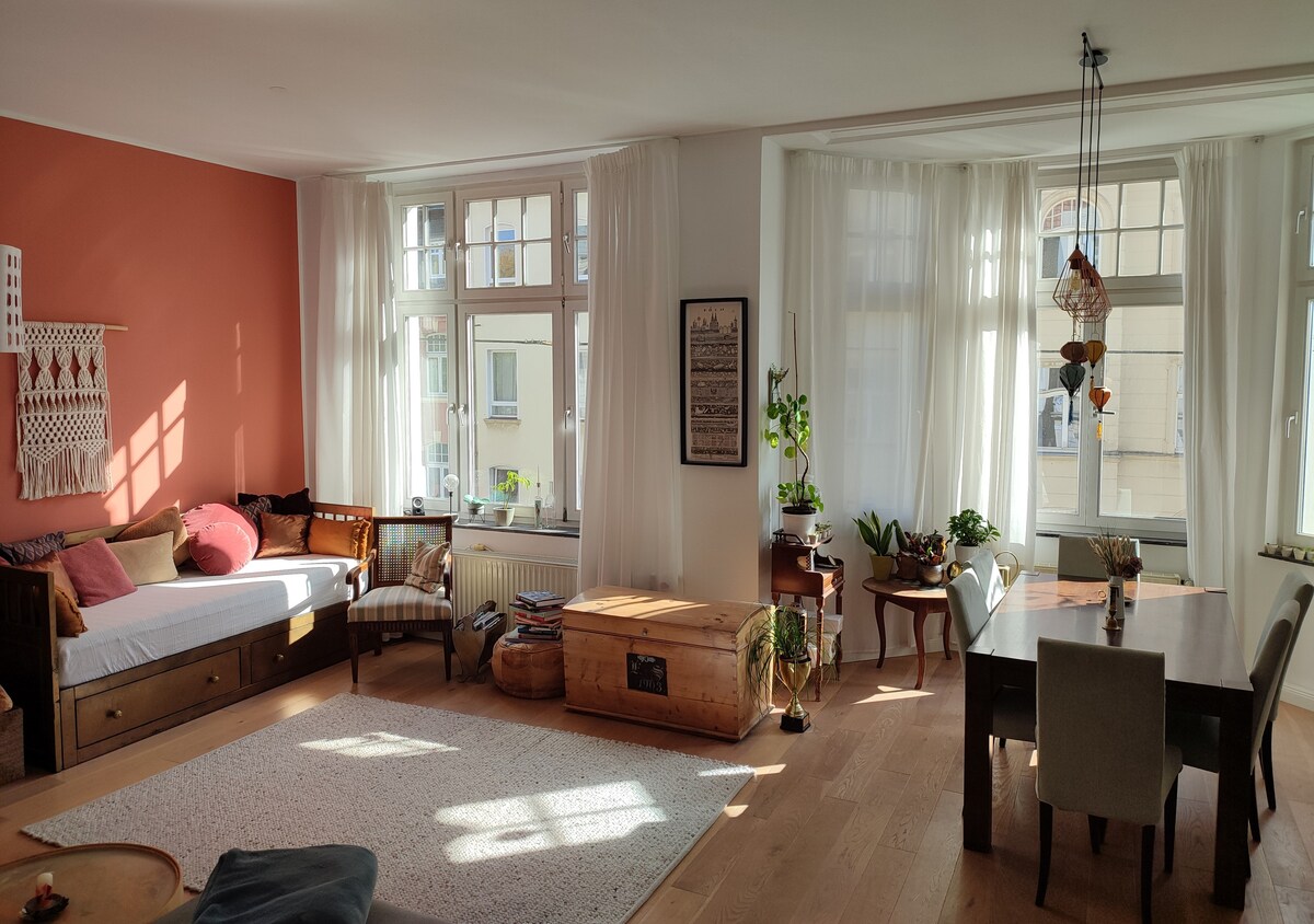 A bright living area features large windows framed by sheer white curtains, allowing ample natural light. A cozy seating arrangement includes a daybed and a wooden chest, complemented by a dining table surrounded by chairs. Decorative plants add a touch of greenery.