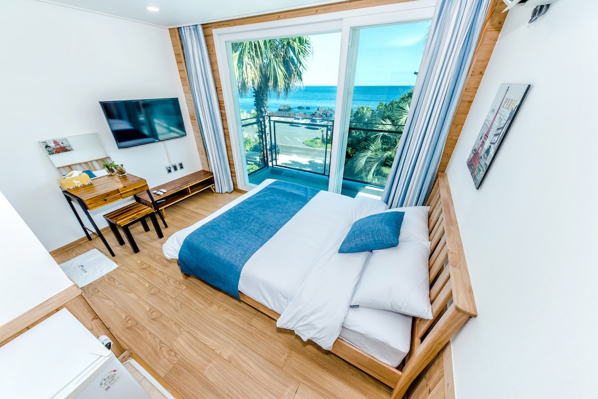 A well-lit bedroom features a spacious bed with white linens and a blue accent blanket. A flat-screen TV is mounted on the wall opposite. A small wooden table and benches provide seating, while large windows offer scenic views of the ocean and surrounding landscape.