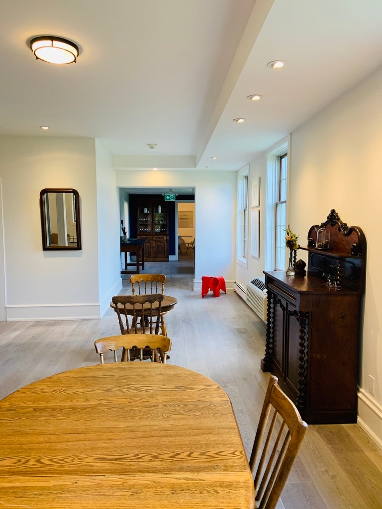 A long, well-lit hallway connects various areas of the space. Light hardwood flooring complements antique wooden furniture, including a round dining table surrounded by wooden chairs. An ornate cabinet and a wall mirror are visible in the background, enhancing the decorative elements.