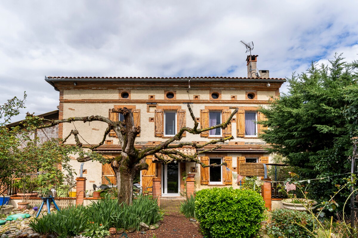 A charming two-story house is surrounded by lush greenery, featuring warm-colored exterior walls and wooden shutters. A welcoming entrance is framed by a blooming garden and carefully maintained shrubs, complemented by a large tree that extends its branches towards the sky.