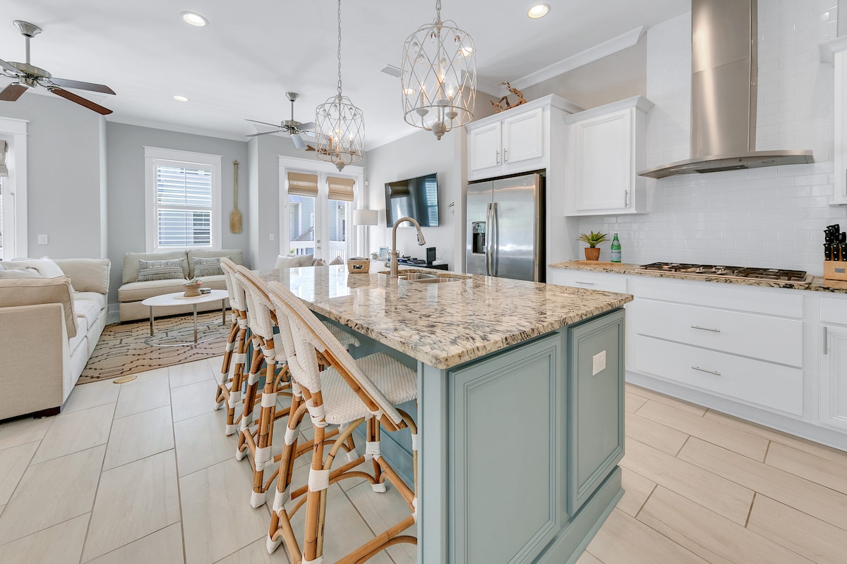 The kitchen features a large island with seating for six, complemented by stylish pendant lights. White cabinetry and modern appliances create a sleek look. Natural light fills the open space, which connects seamlessly to the adjoining living area.