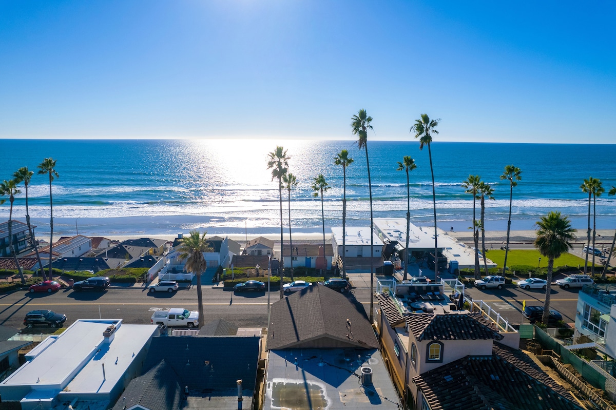 An expansive view of the ocean is captured under a clear blue sky, shimmering under bright sunlight. Tall palm trees line the coast, contrasting with the tranquil beach and waves. The vibrant water reflects the light, creating a serene coastal atmosphere.