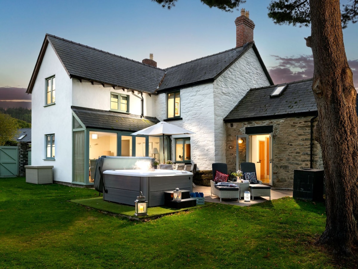 The exterior of a charming two-story farmhouse is illuminated by evening light. A spacious hot tub sits adjacent to the patio, surrounded by well-kept lawn and outdoor seating. The building features a mix of smooth white walls and dark roof tiles, with large glass doors opening to the interior.