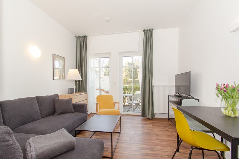 A welcoming living area features a spacious dark couch and a bright yellow chair. A flat-screen TV is positioned on a table across from a dining area with a small, elegant table and chairs. Natural light enters through double doors leading to a terrace.