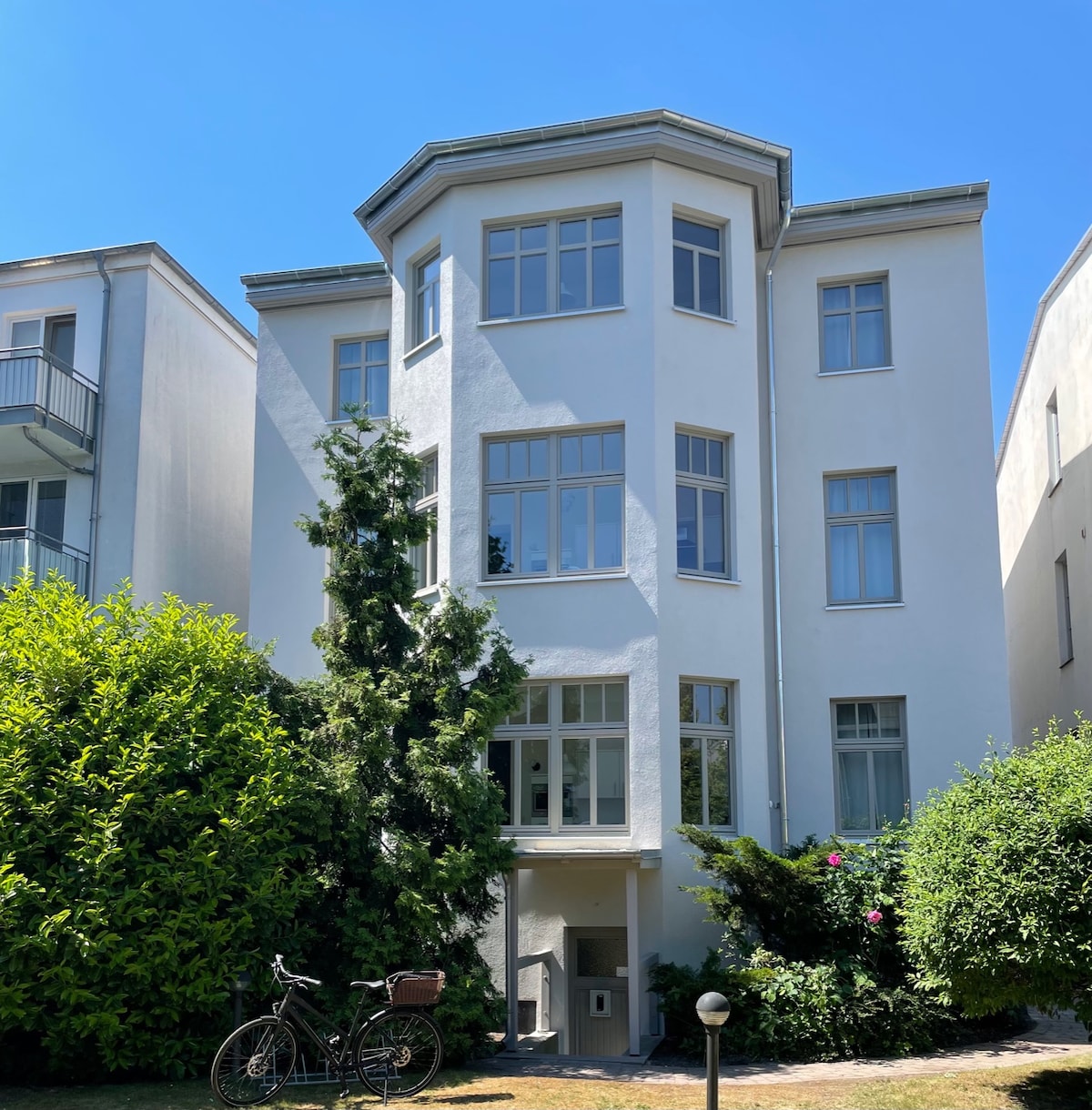 The exterior of a three-story building is displayed, featuring large windows and a light-colored facade. Lush greenery surrounds the structure, alongside a bicycle parked near the entrance. A clear blue sky creates a bright atmosphere.