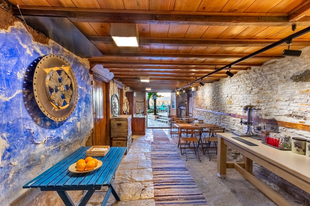 A rustic indoor space features textured stone walls and wooden beams across the ceiling. A blue table with oranges stands prominently, while wooden dining tables are arranged along the sides. The room leads to an outdoor area, visible through the open doorway at the far end.