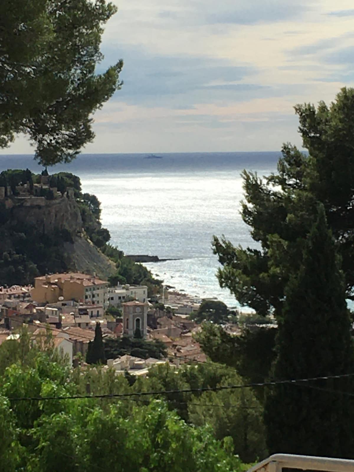A panoramic view captures the shimmering sea and coastal landscape. The distant horizon features a boat gliding over the water, while the charming town sprawls along the shore, framed by lush greenery and the silhouette of hills.