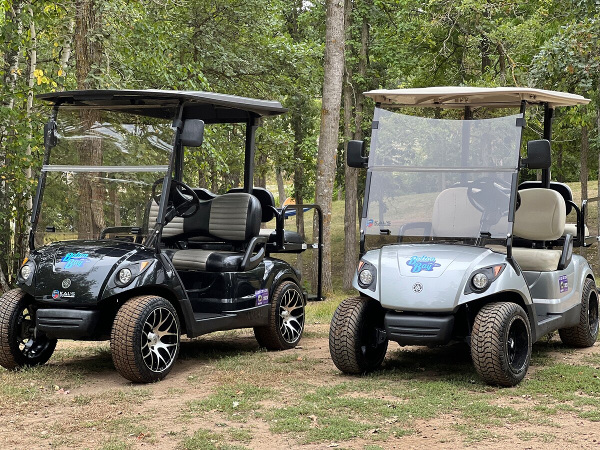 Two golf carts are parked on a grassy area surrounded by trees. One cart has a black exterior with black seats, while the other features a light gray exterior with cream-colored seats. Both carts are designed to seat four passengers and are street-legal.