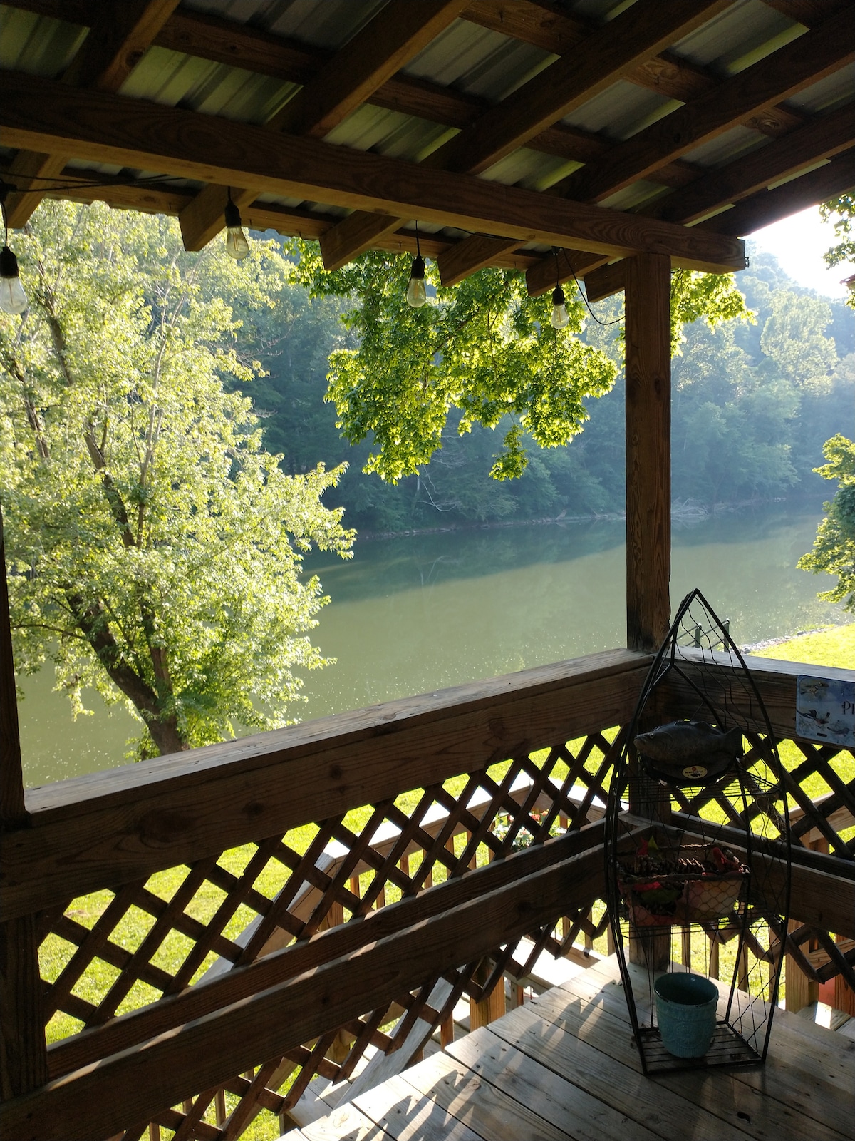 A wooden covered deck is shown, featuring a view of the Greenbrier River surrounded by lush trees. Soft morning light filters through the foliage, casting gentle shadows on the deck floor. A decorative metal stand is visible, enhancing the outdoor space.
