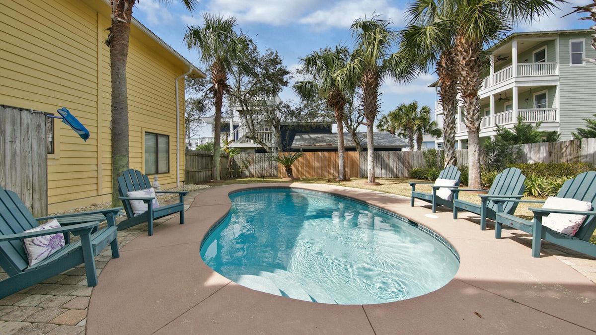 A private pool is surrounded by palm trees and a spacious patio area, featuring several blue lounge chairs for relaxation. Sunlight filters through the trees, casting gentle shadows on the water's surface, creating a serene backdrop for outdoor enjoyment.