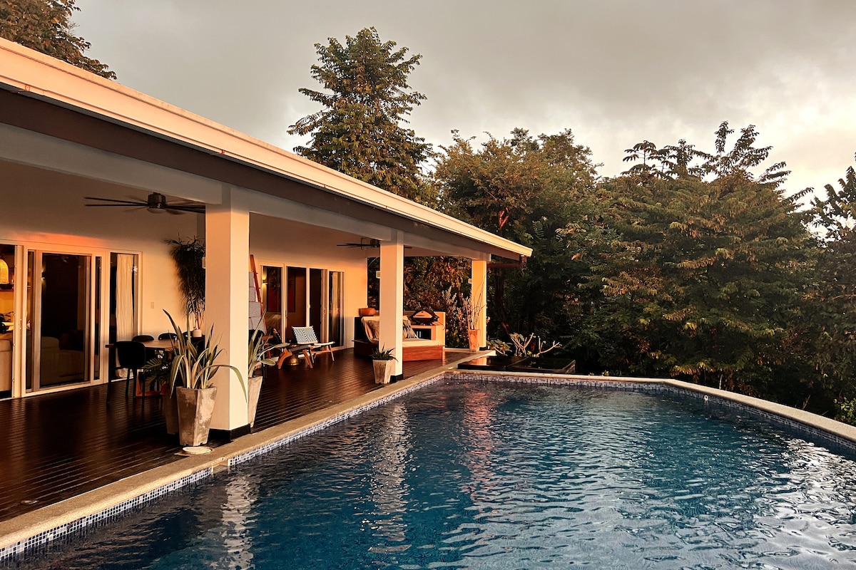The image captures a modern villa overlooking a tranquil pool that reflects the surrounding trees. Large glass doors provide a connection to the outdoor space, with stylish seating arrangements visible near the edge of the pool. Soft lighting enhances the serene atmosphere.