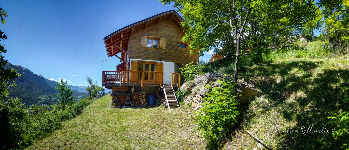 The wooden chalet is nestled on a grassy hillside, showcasing a spacious balcony with wooden railings. Surrounding greenery and distant mountain views enhance the tranquil setting. The structure features large windows, allowing natural light to brighten the interior.