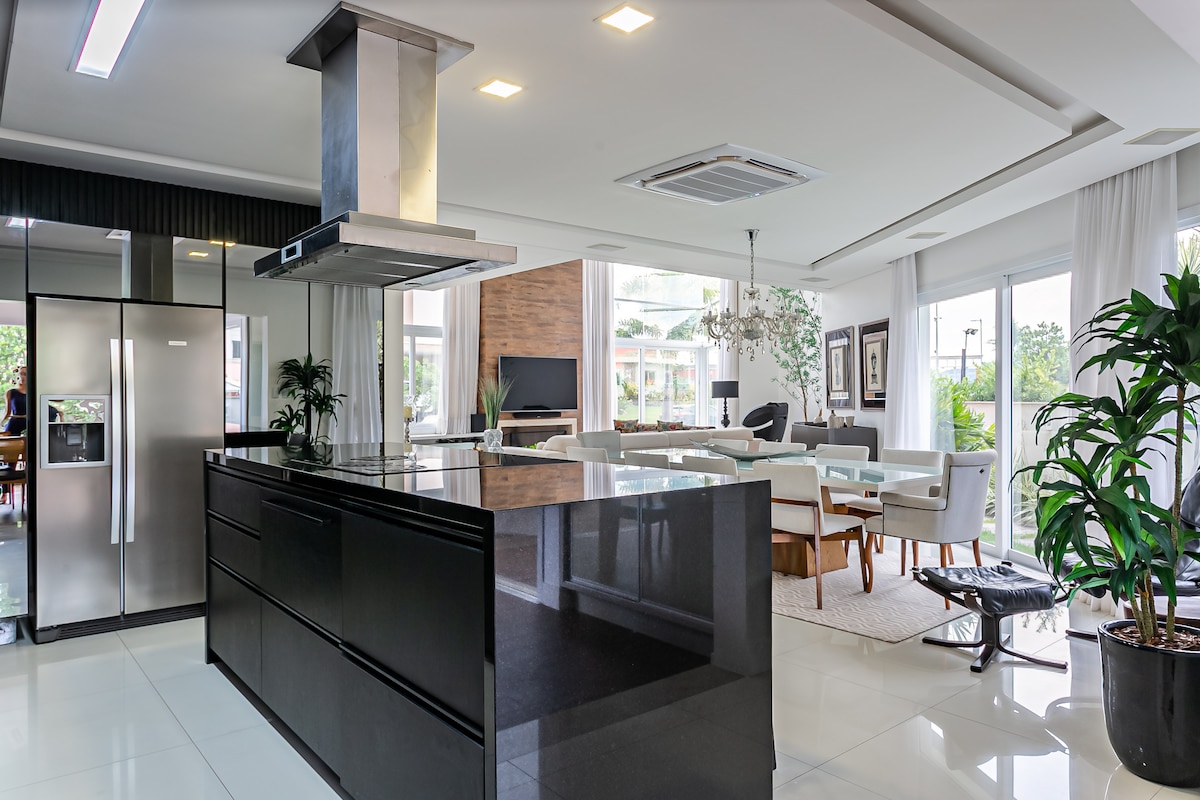 A modern kitchen and living area are presented in an open-concept layout. A large dark countertop with recessed lighting contrasts against sleek cabinetry and stainless-steel appliances. Natural light floods the space through large windows, enhancing the greenery of potted plants positioned around the room.