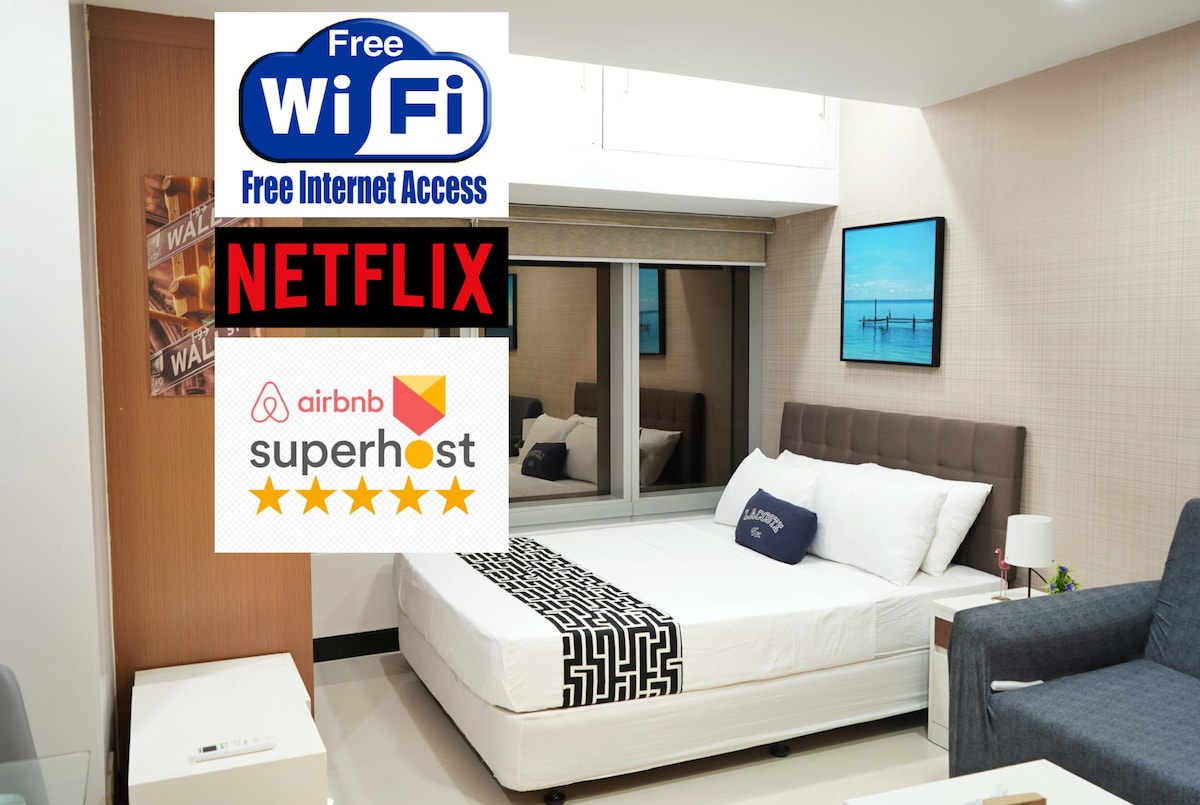 A well-lit modern space features a queen bed with a simple black and white design. A gray sofa is situated nearby. Large windows with blackout blinds allow for light control. Wall art and a TV are also visible, indicating entertainment options.