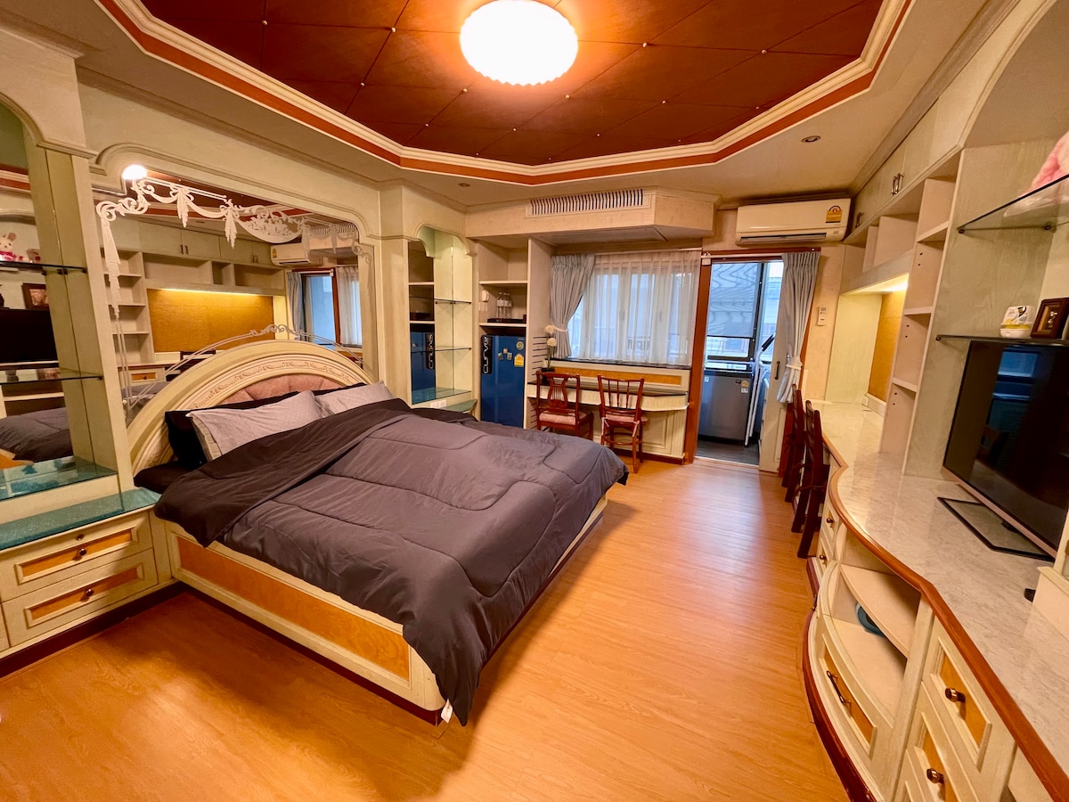 A spacious studio room features a comfortable king-size bed covered with dark bedding. Light streams in through large windows, illuminating built-in shelves and a flat-screen television. A dining table is positioned near the kitchenette, showcasing a convenient layout for living and dining.