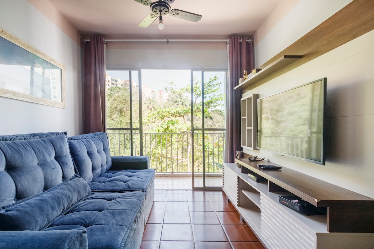 A comfortable living room is highlighted by a plush sectional sofa, facing a mounted television. Large sliding glass doors allow natural light to fill the space, revealing views of greenery outside. Neutral walls and wooden shelves add to the inviting atmosphere.