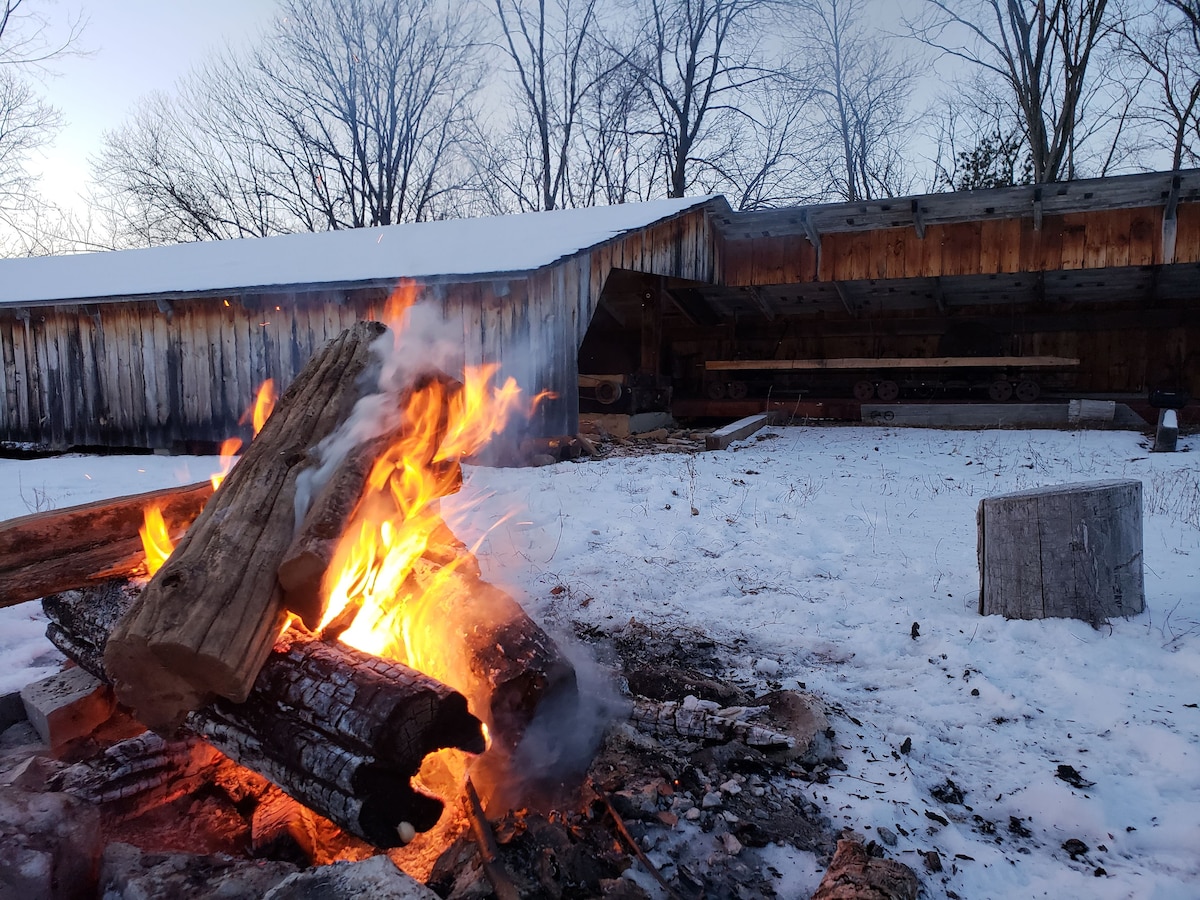 A warm campfire crackles amidst the snowy landscape, with logs burning gently. The restored historic sawmill stands in the background, its wooden structure partially covered in snow. Leafless trees frame the scene, creating a serene winter atmosphere.