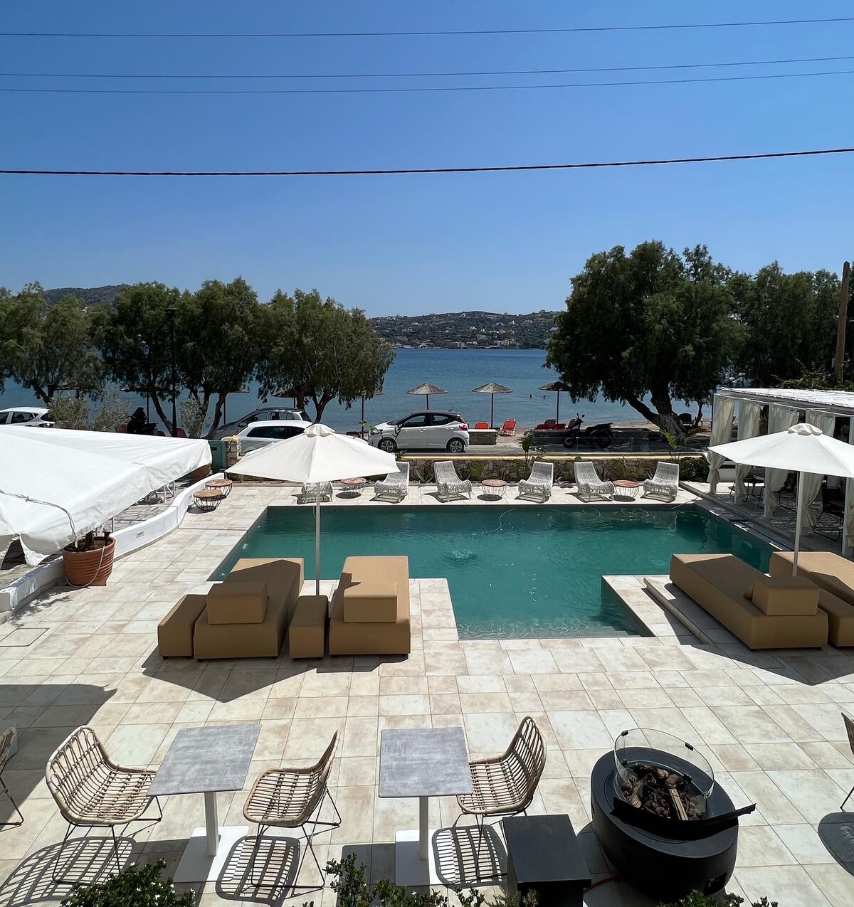 A spacious outdoor area surrounds a tranquil pool with comfortable lounge seating. Umbrellas provide shade, while the serene sea view is visible in the background. The tiled patio features arranged tables and chairs, creating an inviting space for relaxation.