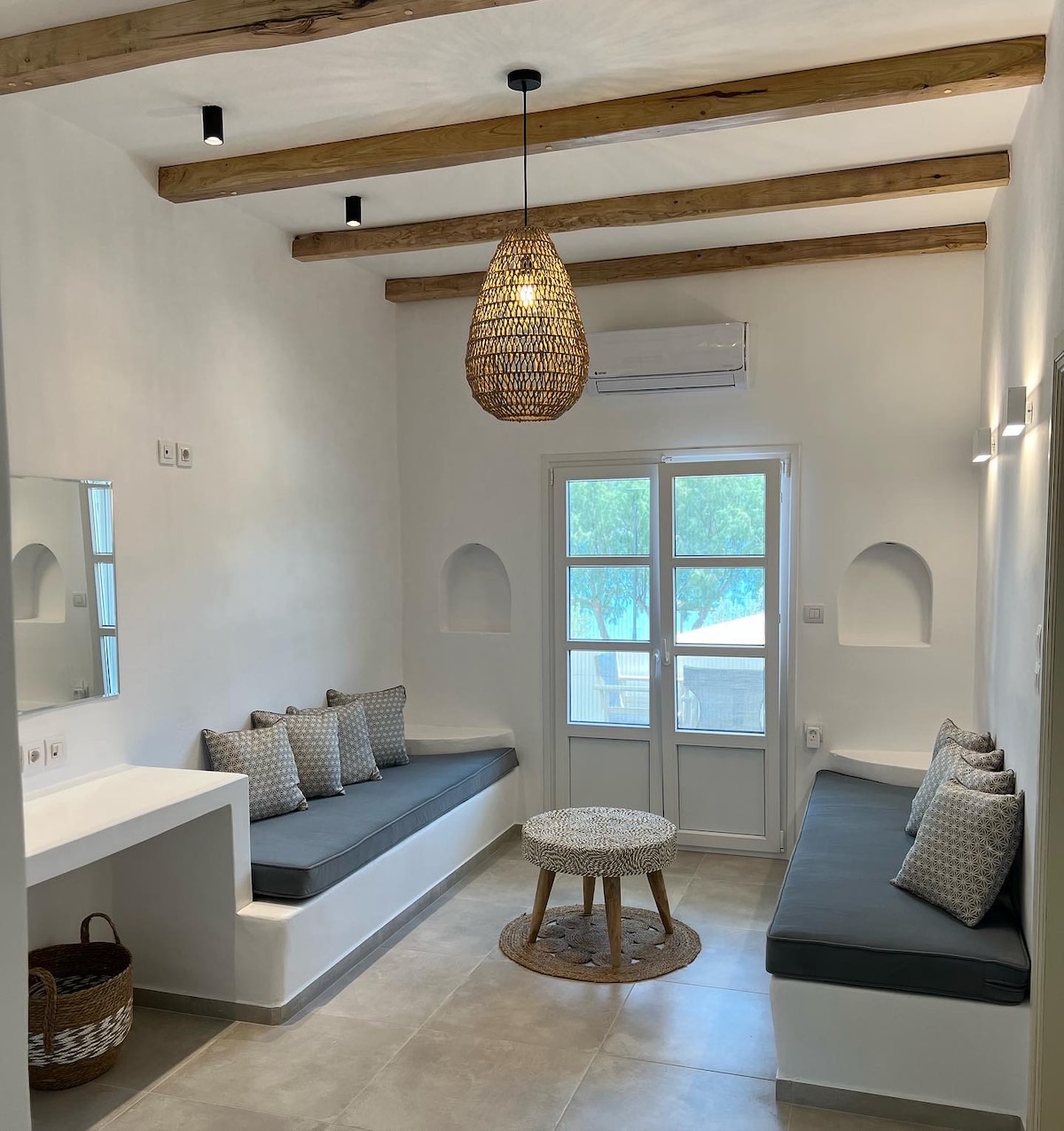 A minimalist sitting area features built-in benches adorned with decorative cushions. A round wooden table is placed at the center, and natural light enters through a door with multiple panes. Wooden beams add warmth to the white walls, creating an inviting space.