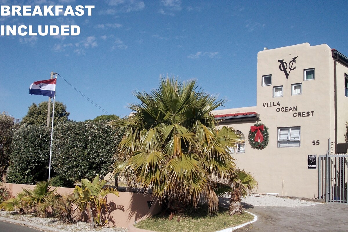 The exterior of the guesthouse features a beige building adorned with a decorative wreath. A tall palm tree is visible in the foreground, alongside a flagpole displaying a flag. The street address '55' is prominently marked on the wall.