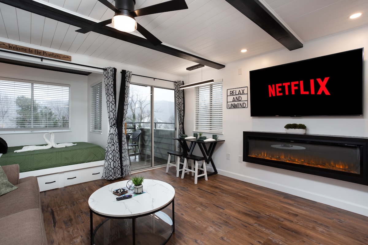 A modern living space is presented, featuring a large electric fireplace below a 65" smart TV displaying Netflix. A cozy couch and a round coffee table contribute to the relaxed atmosphere. A private balcony is accessible through glass doors, framed by light curtains.
