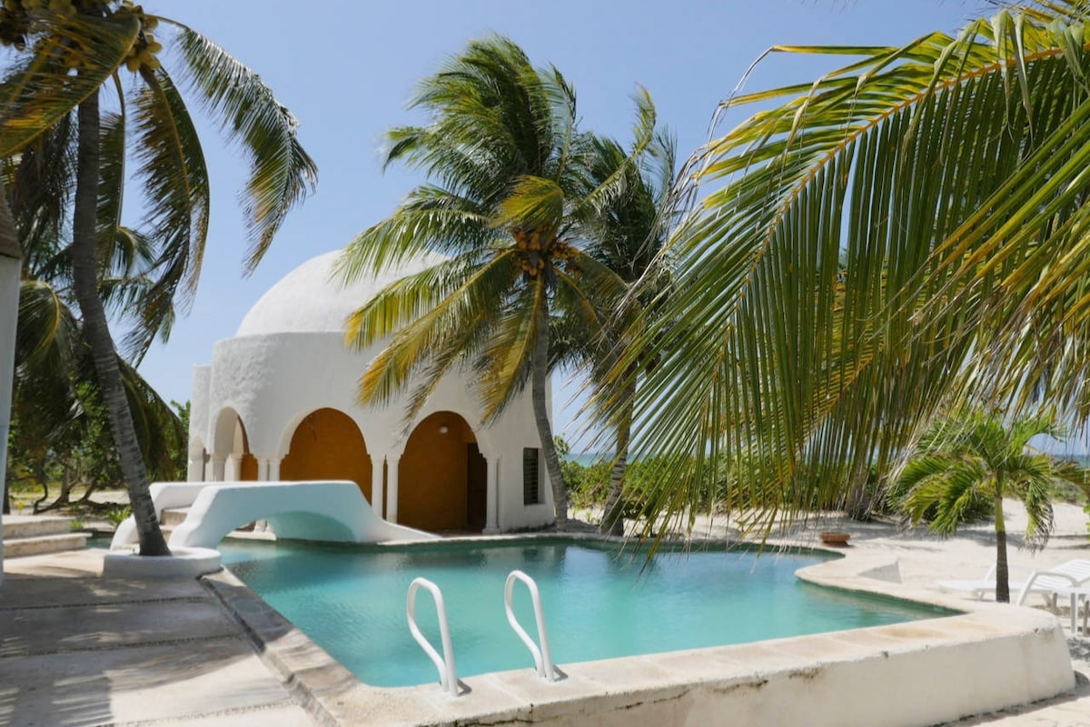 A white villa with a distinctive dome shape is positioned beside a clear swimming pool, surrounded by palm trees. The pool deck features a set of steps leading into the water, creating an inviting outdoor space. Bright blue skies enhance the serene atmosphere.