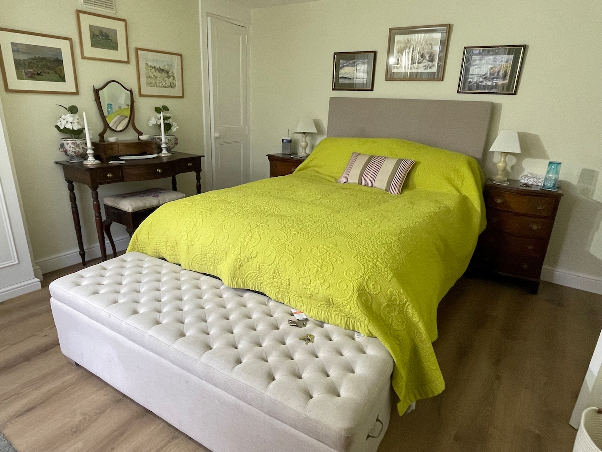 A bright bedroom is furnished with a spacious bed covered in a vibrant yellow quilt. Bedside tables with lamps flank the bed, and a tufted bench is positioned at the foot. Framed artworks decorate the walls, enhancing the room's charm.