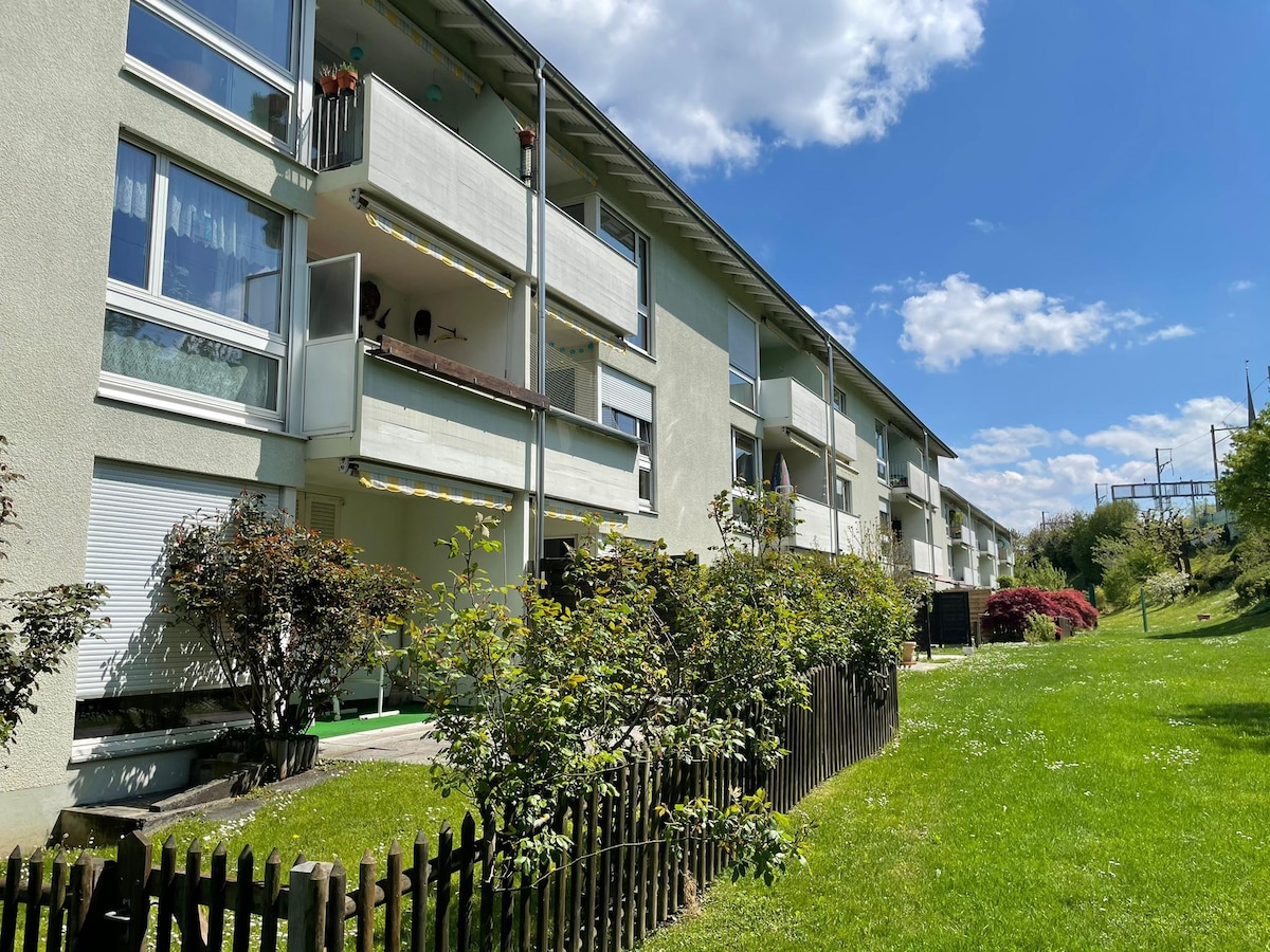 A three-story apartment building is depicted, featuring several balconies and a manicured lawn. Green bushes border the grassy area, contributing to the tranquil atmosphere. The blue sky above is dotted with a few clouds, creating a sense of openness and light.