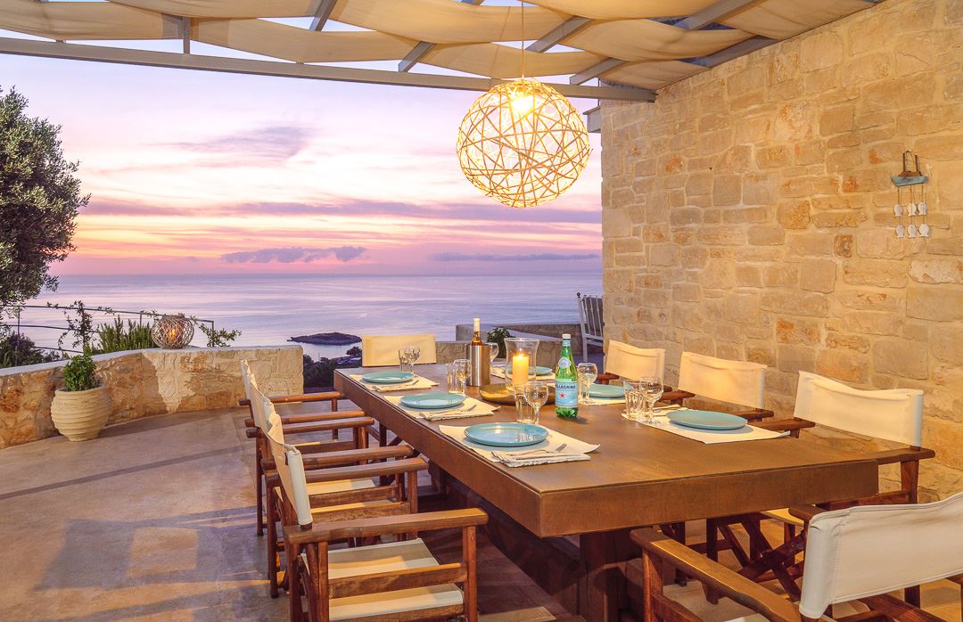 An outdoor dining area features a large wooden table surrounded by comfortable chairs. A stylish light fixture hangs above, while the vivid colors of a sunset reflect off the tranquil sea in the background, enhancing the scenic coastal view.