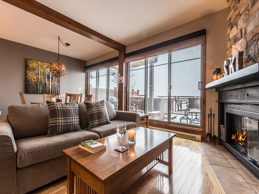 A comfortable living area features a plush sofa adorned with decorative pillows. A coffee table is positioned in front of a stone fireplace, and large windows provide views of the outdoor terrace. Natural light fills the space, highlighting wooden accents throughout.