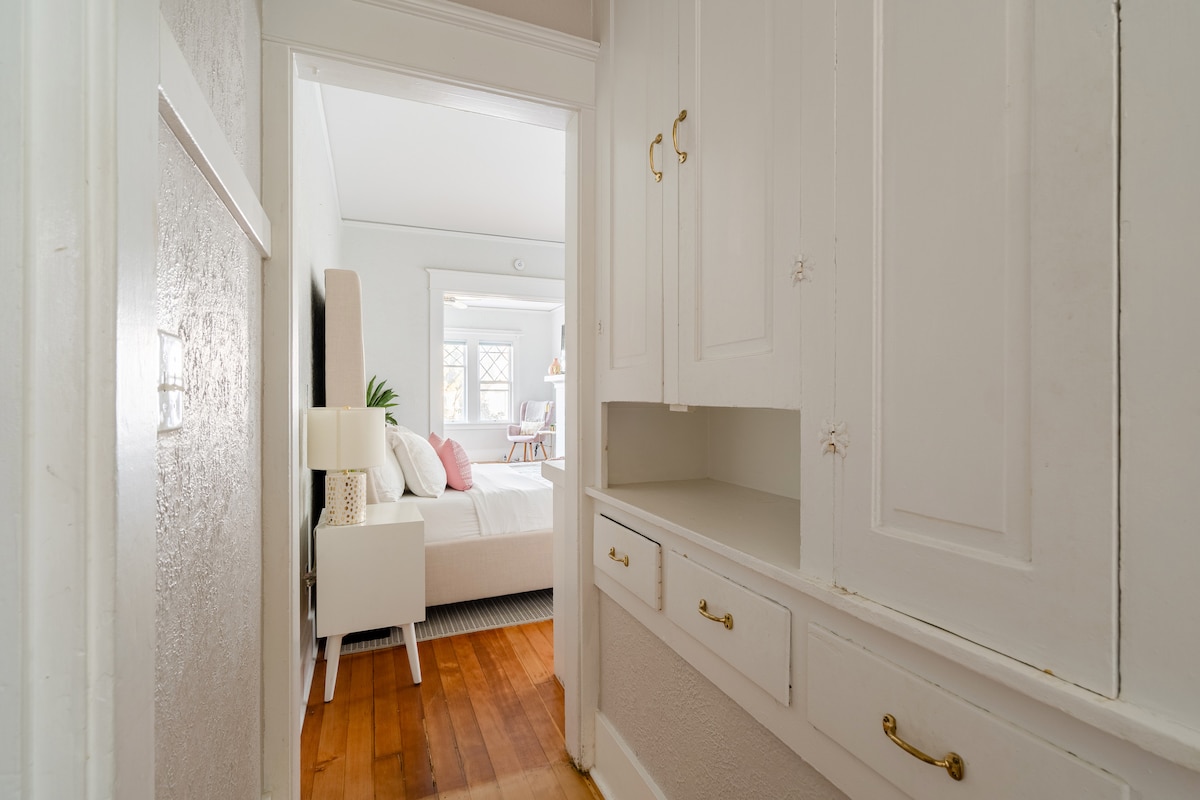 A hallway leads to a bright bedroom, displaying white cabinetry with brass handles. The warm wooden floor contrasts with the light walls, while the bedroom beyond features a spacious bed with soft linens and warm natural light streaming through windows.