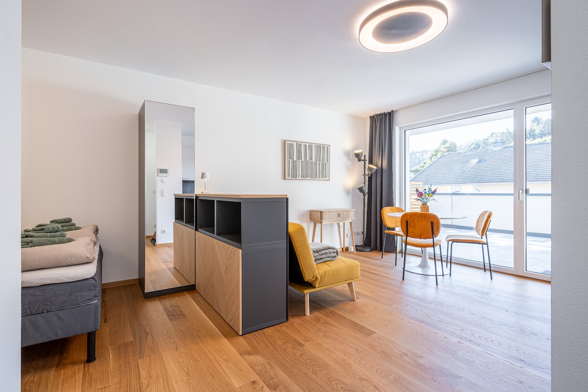 A spacious studio apartment is shown, featuring a comfortable bed placed next to a large mirror. A cozy chair in warm tones complements a dining area with four chairs. Natural light enters through a large sliding door that opens to a terrace.