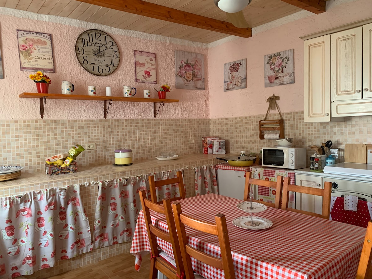 The kitchen features light-colored cabinetry and a checkered tablecloth covering a wooden dining table. Shelves display decorative items and a large clock hangs on the wall. The space includes essential appliances and a patterned curtain that complements the warm, inviting atmosphere.