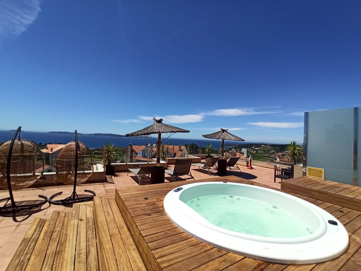 A wooden deck features a hot tub surrounded by sun loungers and umbrellas. In the background, a panoramic view of the sea and distant islands is visible under a clear blue sky.