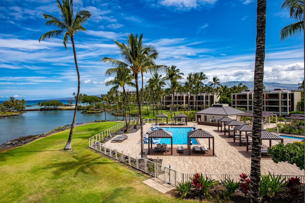 A serene view of the pool area features elegant cabanas and lounge chairs surrounding a large heated pool. Lush green landscaping and palm trees frame the scene, while a glimpse of the ocean and Hawaiian fishponds can be seen in the background.