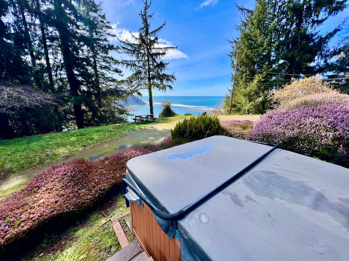 A private hot tub is displayed in the foreground, offering serene views of the ocean beyond. Lush greenery and flowering bushes surround the area, while tall trees provide shade. A picnic area is visible in the distance, enhancing the natural setting.