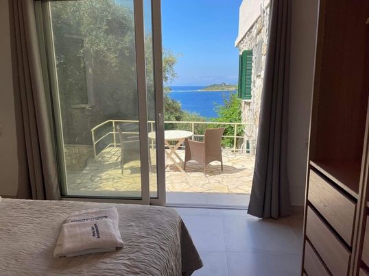 A serene bedroom features floor-to-ceiling glass doors that open to a private terrace. Outside, a small table and two chairs are positioned, with a view of the sea and surrounding greenery. The room includes light-colored furnishings and a neatly made bed.