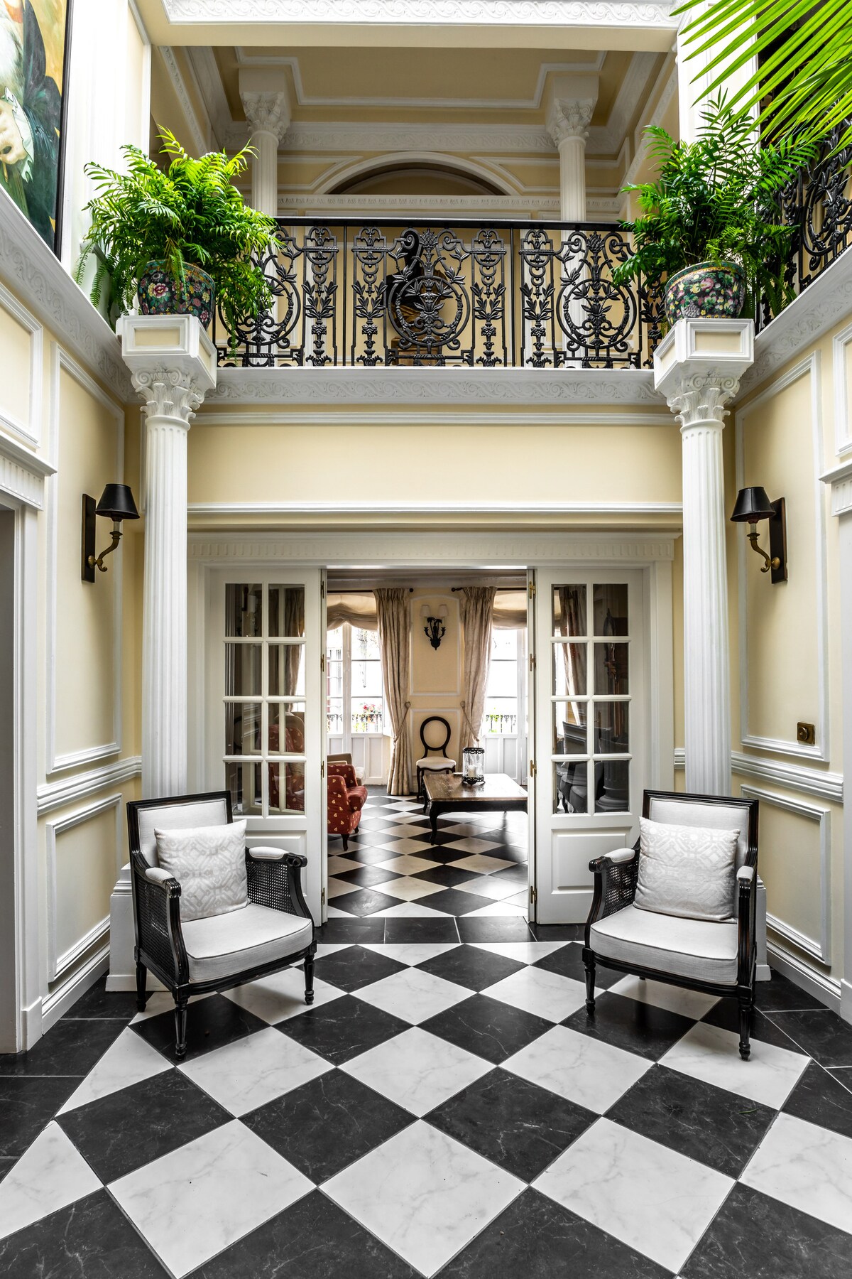 The entrance hall features a striking black and white checkered floor, complemented by elegant wall mouldings and tall ceilings. Two armchairs are positioned near glass-paneled doors, while a wrought-iron balcony overlooks the area. Lush greenery adds a vibrant touch to the upper level.