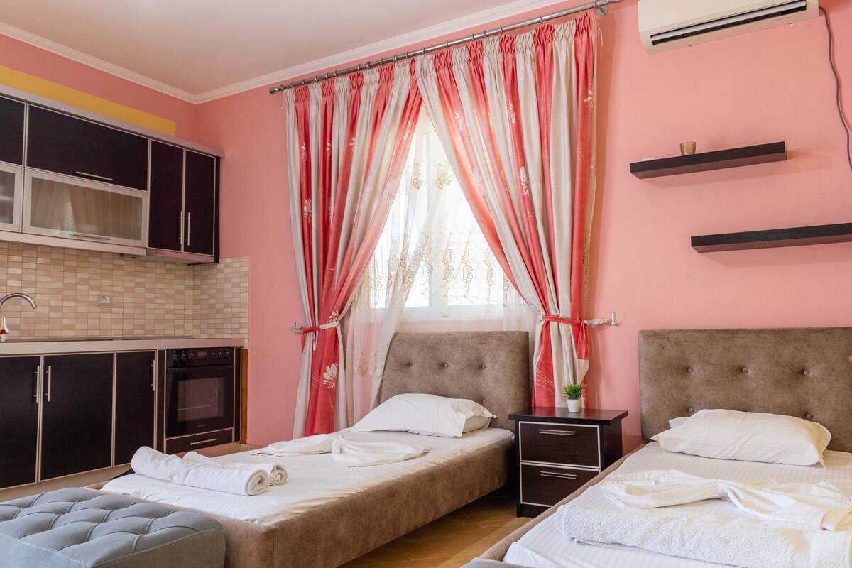 A bright room features two spacious beds with crisp linens, positioned beside a dark wooden nightstand. A kitchenette with modern cabinetry is visible, and large windows are adorned with flowing sheer curtains, allowing natural light to brighten the pink walls.