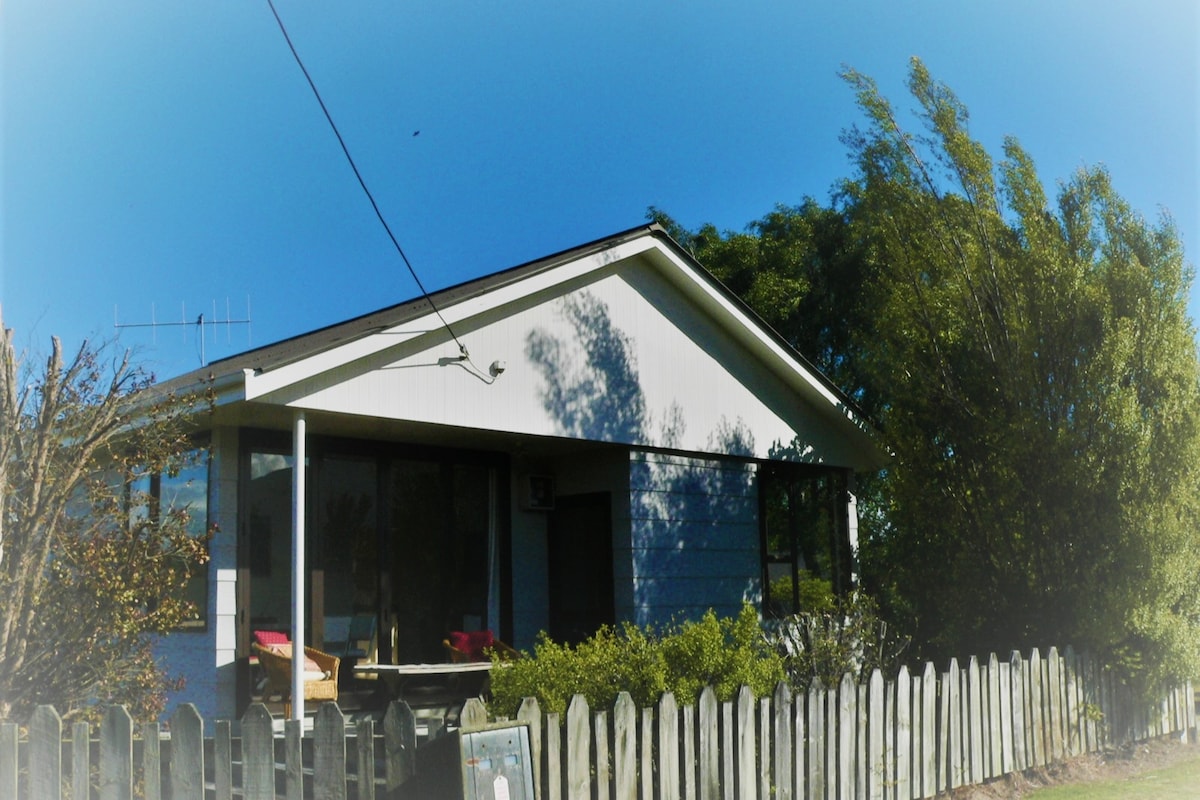 The exterior of the home is depicted, featuring a light-coloured façade and large windows that allow natural light to fill the interior. A wooden fence borders the front yard, with surrounding greenery and a clear blue sky overhead.
