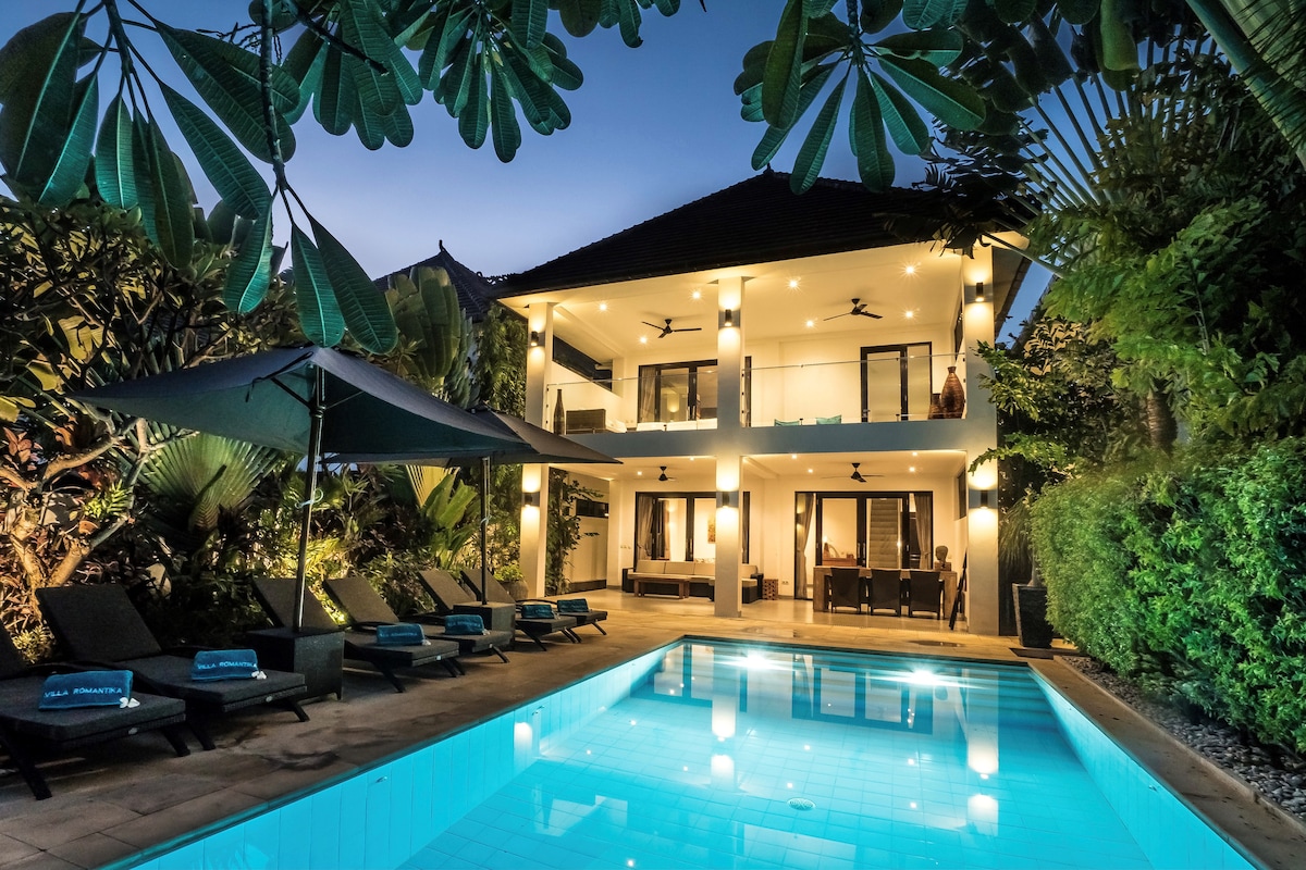 The villa is illuminated at dusk, showcasing a private pool surrounded by tropical foliage. Comfortable lounge chairs are positioned by the pool, and an outdoor dining area is visible on the terrace. The two-story structure features multiple glass doors, enhancing the connection between indoor and outdoor spaces.