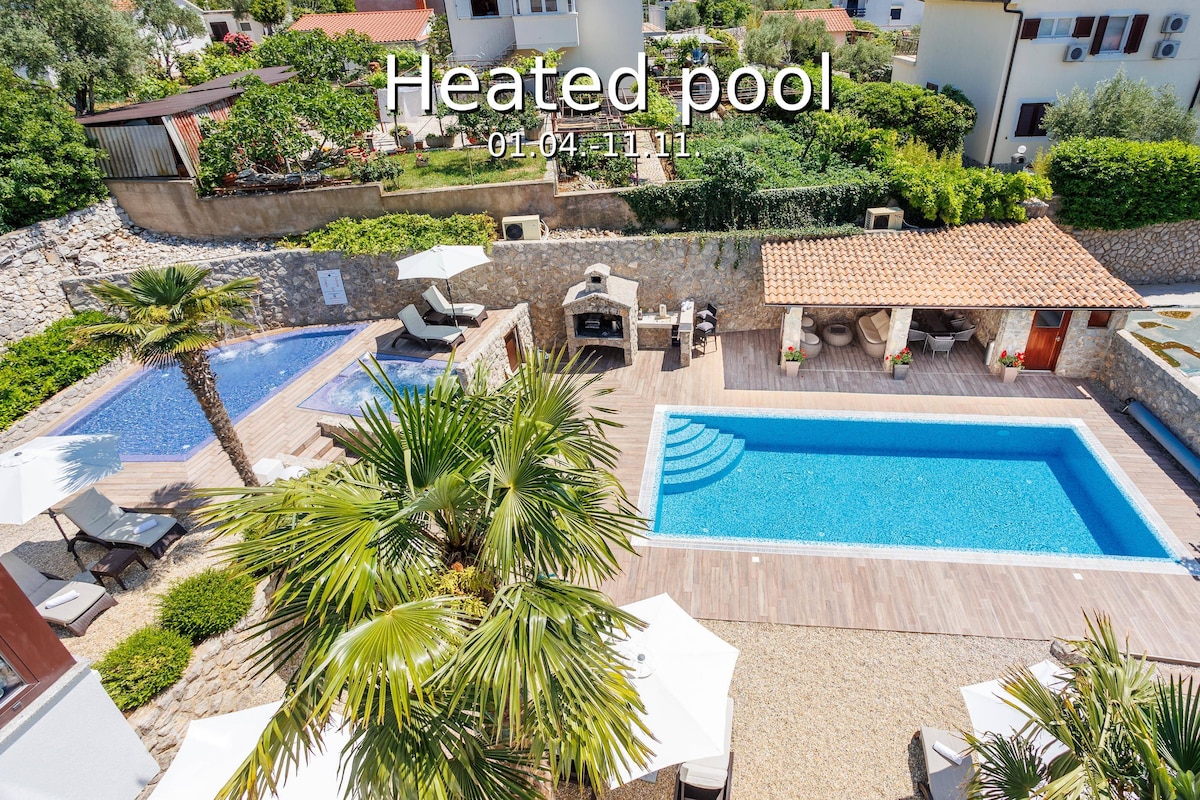 A heated outdoor pool is surrounded by a wooden deck, complemented by lounge chairs and shaded areas. Lush greenery and palm trees provide a serene atmosphere. A separate area features a barbecue setup alongside a second pool, enhancing the outdoor space for relaxation and leisure.