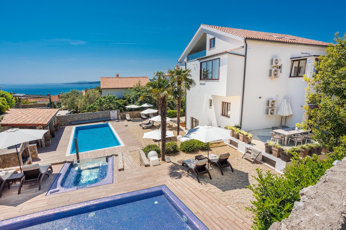 A spacious outdoor area features a heated pool and a separate hot tub, surrounded by lounge chairs and umbrellas. The modern building is complemented by lush greenery and offers views of the sea. A dining area is positioned nearby for outdoor meals.